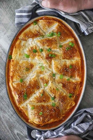 An overhead view of sloppy Joe casserole in an oval baking dish that is topped with browned puff pastry and garnished with sesame seeds and chopped scallions.