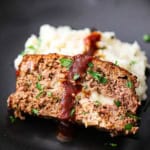 A close-up view of a slice of cheese-stuffed smoked meatloaf resting on a pile of mashed potatoes on a black plate.