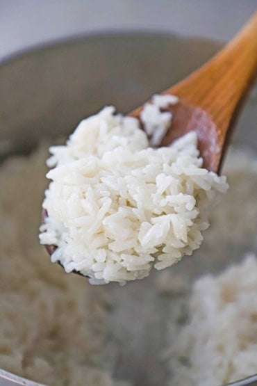 A close-up view of a wooden spoon that is holding a serving of easy coconut rice over a saucepan that is filled with the same rice.