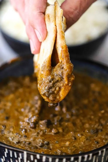 A straight-on view of a person holding a folded piece of naan bread that has been plunged into a bowl of homemade dal makhani and is being held above the bowl with lentils dripping from it.