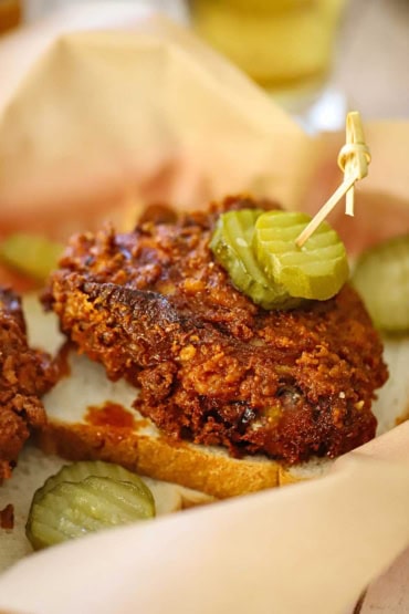 A close-up view of a piece of Nashville hot chicken sitting on a slice of white bread with two dill pickle slices attached to the chicken with a toothpick.