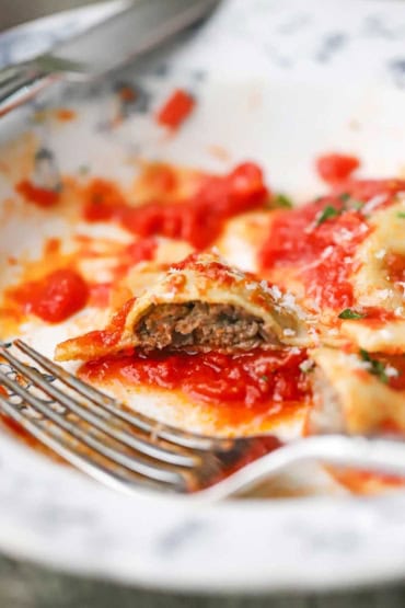 A close-up view of homemade beef ravioli that is on a white plate with a bite take from one ravioli with the beef interior revealed.