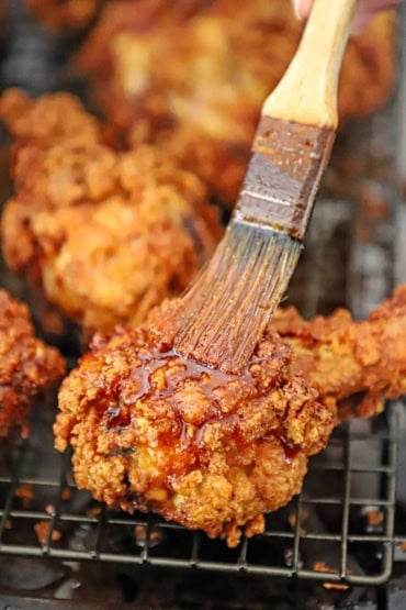 A person using a small brush to apply Nashville hot oil to a piece of Nashville fried chicken which is crispy leg on a baking rack.