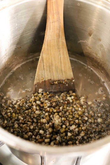 A view into a large stock pot that is filled with black lentils that have been simmering for several hours in a water with a wooden spatula inserted into the side.