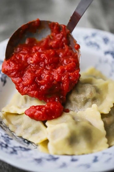 A ladle being used to transfer a tomato sauce onto a pile of cooked homemade beef ravioli in an antique bowl.