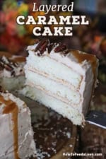 A person using a large knife turned sideways to lift a slice of caramel layer cake from a wooden serving platter.