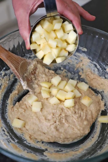 A person transferring cubed chunks of fresh apple into a large glass bowl filled with an apple fritters batter.