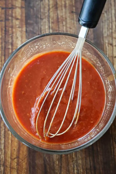 An overhead view of a glass bowl filled with homemade cocktail sauce with a large whisk in the middle of it.