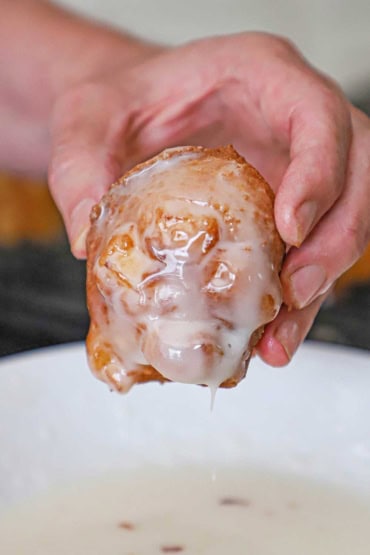 A person lifting a freshly fried homemade apple fritter from a bowl filled with cider glaze with some of the glaze dripping from the fritter.