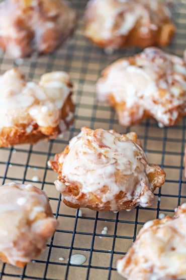 A close-up view of a collection of homemade apple fritters resting on a baking rack with drips of cider glaze dripping from the freshly fried fritters.