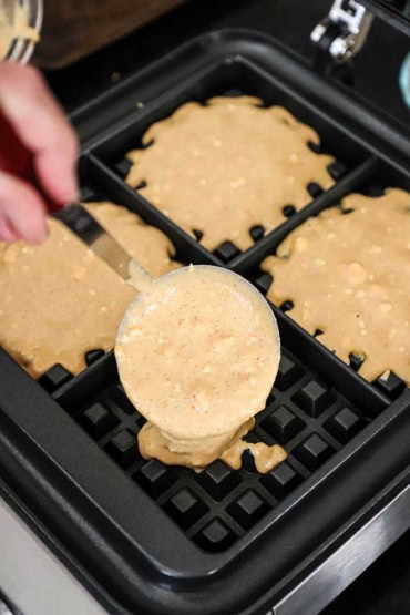 A person using a metal measuring cup to transfer pumpkin waffle batter into a four chamber waffle maker.