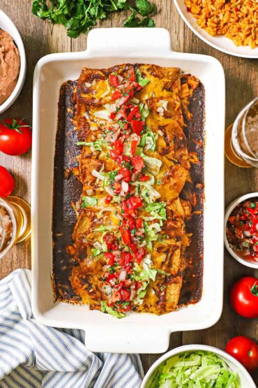 An overhead view of a rectangular white baking dish filled with leftover turkey enchiladas in an enchilada red sauce and topped with chopped lettuce and tomatoes.