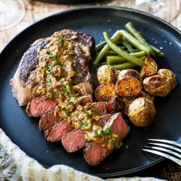 A straight-on view of a reverse-sear ribeye steak with gorgonzola on a black dinner plate alongside a helping of roasted potatoes and steamed green beans.