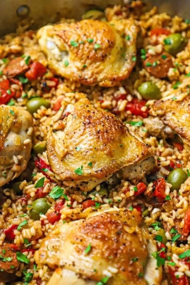 A close-up view of Spanish chicken and rice in a skillet with strips of roasted bell peppers and green olives scattered around the chicken in the rice.