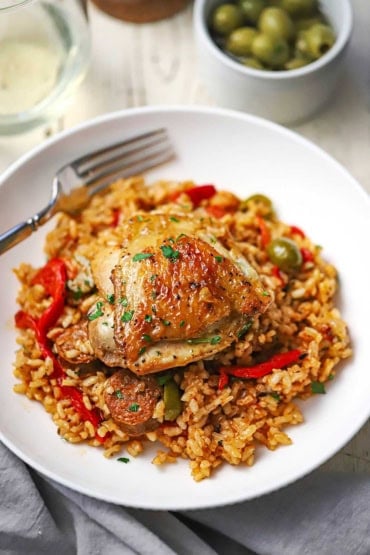 A close-up view of a serving of Spanish chicken and rice in a serving bowl with one seared chicken thigh resting on the colorful rice with slices of Spanish chorizo visible.