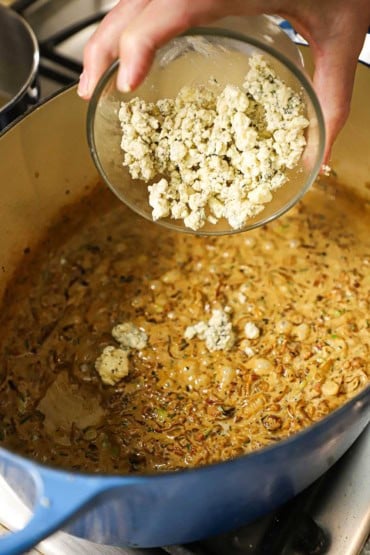 A person transferring crumbled Gorgonzola cheese from a small glass bowl into an oval Dutch oven that is filled with a simmering caramelized onion and cream sauce.