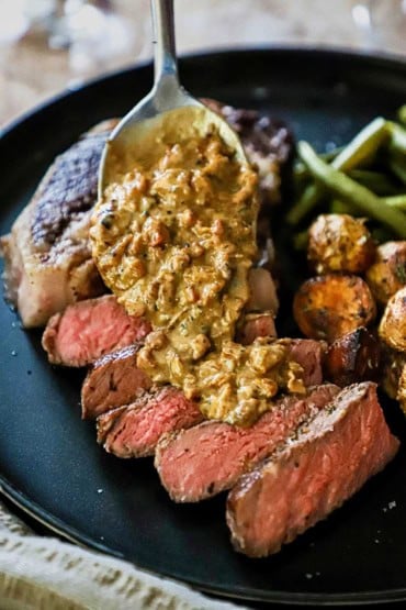 A person using a silver spoon to drizzle a Gorgonzola sauce over a sliced ribeye steak on a black plate also holding roasted potatoes and green beans.