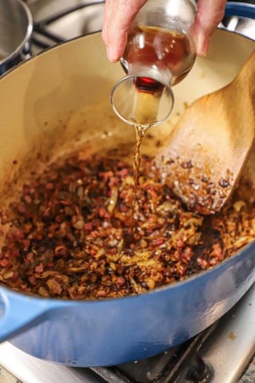 A person pouring Sherry from a small wine carafe into an oval Dutch oven filled with caramelized onions and crispy cubed pancetta.