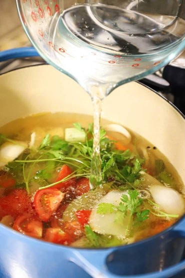 A person pouring water from a large glass measuring cup into an oval Dutch oven filled with chopped vegetables and fresh parsley.