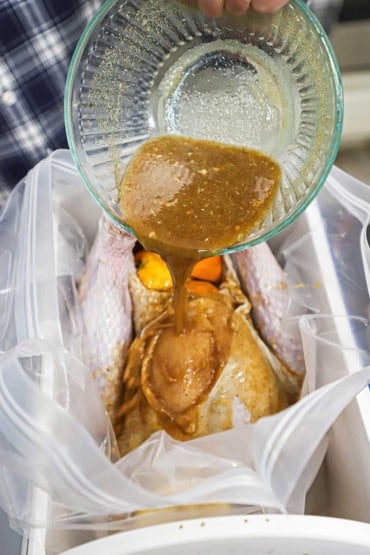 A person pouring a orange juice and maple marinade from a glass bow over an uncooked turkey that is snuggled into a brine bag in an upright cooler.