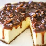 A close-up view of a cheesecake topped with a pecan pie sauce with a slice of cake missing.