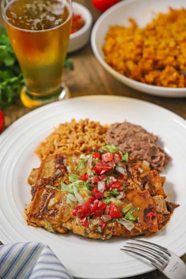 A straight-on view of a white dinner plate filled with two leftover turkey enchiladas next to a serving of refried beans and Mexican rice with a glass of beer in the background.