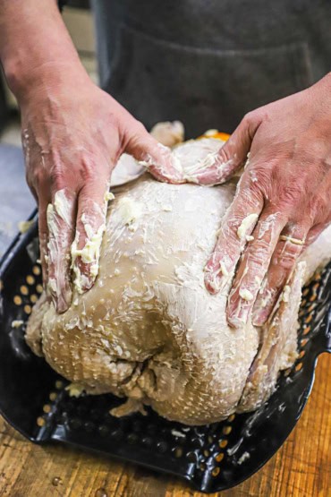 A person using his hands to rub softened butter all over the exterior of an uncooked whole turkey resting on a roasting rack.