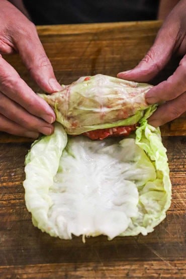 A person using his two hands to roll an uncooked beef and rice mixture within a cabbage leaf on a wooden cutting board.