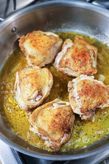 A straight-on view of five skin-on, bone-in chicken thighs that have been seared in a large skillet in olive oil and the skin visible and is browned and crispy.