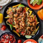 An overhead view of a large fajita skillet filled with a serving of shrimp fajitas with bowls of all the fixings nearby including pice de gallo, lime wedges, and crumbled Cotija cheese.