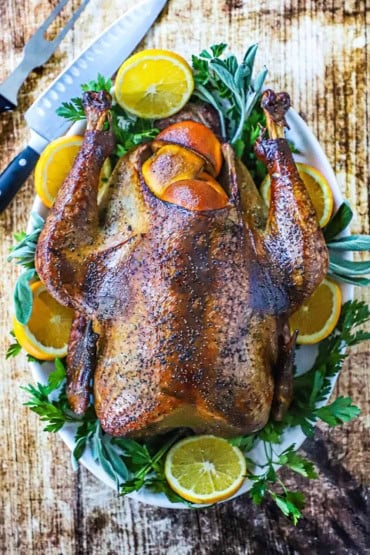 An overhead view of a whole smoked turkey that is resting on a platter of parsley and orange slices and a carving knife nearby.