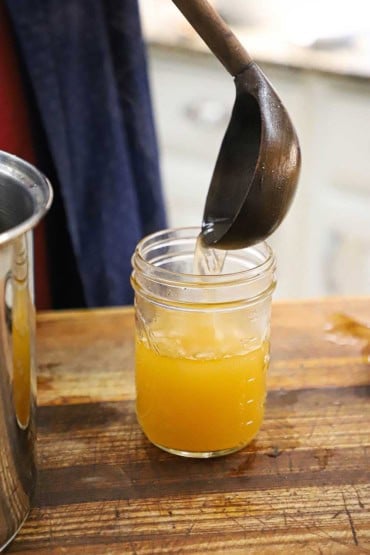 A person using a large wooden ladle to transfer homemade vegetable stock from a stock pan into a glass Mason jar.