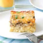 A straight-on view of square piece of a breakfast casserole with a top layer of melty cheese over sausage sitting on a bed of shredded potatoes.