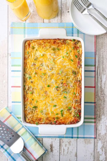 An overhead view of best breakfast casserole in a rectangular white baking dish resting on a colorful place matt next to a glass and carafe of orange juice.