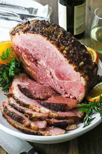 A straight-on view of a half smoked bourbon glazed ham resting on a large white platter with numerous slices cut from the roast on stacked also on the platter.