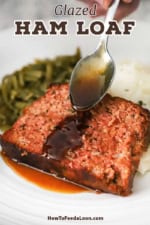 A person using a silver spoon to drizzle a glaze across the top of a glazed ham loaf slice that is resting on a bed of mashed potatoes on a white dinner plate.