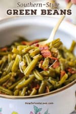 A close-up view of a person raising a large serving spoon of Southern-style green beans from an old-fashioned pot that is filled with the cooked beans.