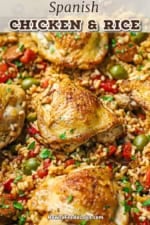 A close-up view of Spanish chicken and rice in a skillet with strips of roasted bell peppers and green olives scattered around the chicken in the rice.