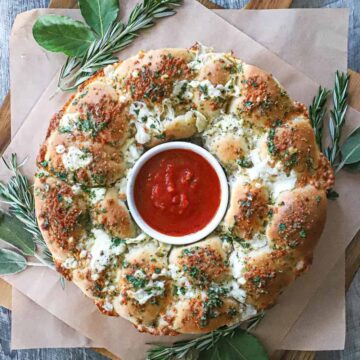 An overhead view a bread wreath with a ramekin in the center filled with marinara sauce and is surround by festive herbs.