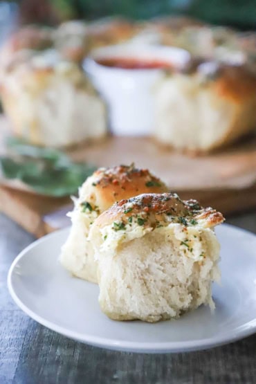 A straight-on view of two homemade cheesy skillet rolls that are sitting on a small white plate with the loaf of rolls in the background.