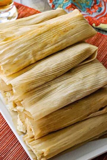 An overhead view of a white rectangular serving platter that is filled with a tall pile of unopened homemade tamales with a glass of beer nearby.