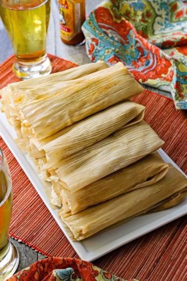 An overhead view of a white rectangular serving platter that is filled with a tall pile of unopened homemade tamales with a glass of beer nearby.