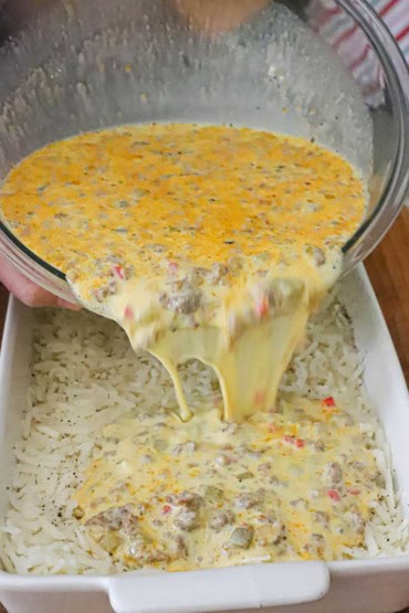 A straight-on view of a person pouring an egg and cream mixture with sausage into a baking dish that is lined with uncooked frozen hashbrowns.