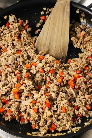 A close-up view of a large black non-stick skillet filled with cooked breakfast sausage with chopped red bell peppers and onion and a wooden spatula inserted in the middle of it.