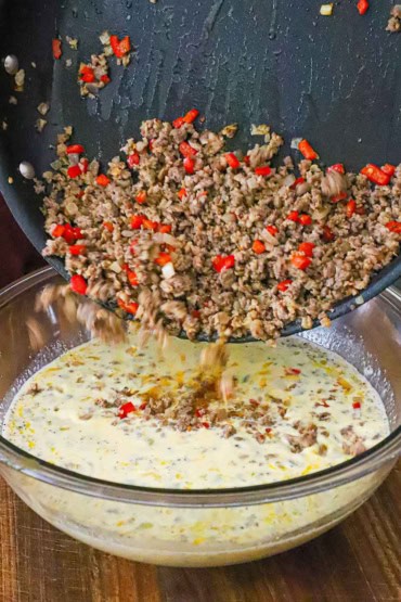 A person dumping cooked sausage and peppers from a skillet into a large glass bowl filled with an egg and cream mixture.