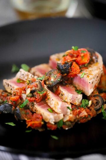 A close-up view of a sliced seared tuna steak that is topped with a Mediterranean sauce of black olives, tomatoes, olives, and garlic.