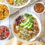 An overhead view of a white bowl holding a serving of turkey burrito bowl topped with slices of avocado, kidney beans, and sour cream and surround by bowls of salsa, pico de galls, and corn.