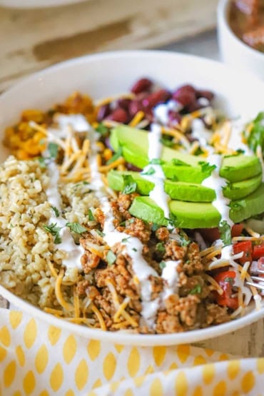 A close-up view of a white shallow bowl holding a turkey burrito bowl topped with slices of fresh avocado and streaks of Mexican crema.