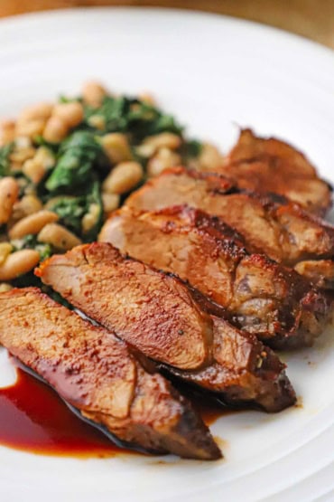 A close-up view of sliced pork tenderloin with a cranberry reduction sauce drizzled on top with a serving of sautéed white beans and spinach next to it.