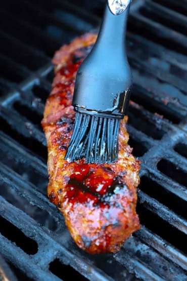 A straight-on view of a black grill brush that is being used to apply a cranberry mop over the top of a pork tenderloin that is being grilled on an outdoor gas grill.